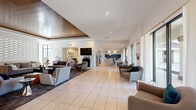 Located at 912 Innovation Way in Altamonte Springs, FL, this single-floor community space offers a generously proportioned, open-concept layout designed with modern sophistication. The entrance greets visitors with soaring ceilings, large-format light tile flooring, and a dramatic oversized art installation set within a recessed wall niche, establishing an immediate sense of grandeur and refinement.

The expansive lounge area is anchored by a striking wood-paneled feature wall housing a flat-screen TV and illuminated display shelving, all beneath a warm wood tray ceiling that adds architectural depth. Comfortable seating arrangements, including plush sofas and contemporary accent chairs, are arranged on layered area rugs, creating an inviting communal atmosphere. A sculptural three-dimensional wall panel allows natural light to filter softly into the seating area.

The combined kitchen and dining area is a functional centerpiece, featuring an extended island with bar seating, sleek white cabinetry, stainless steel appliances, and a textured mosaic tile backsplash. Pendant lighting in globe and floral forms adds a designer touch. The dining nook showcases a live-edge wood table, navy upholstered banquette seating, and elegant chandelier fixtures, with direct access to an outdoor area through floor-to-ceiling glass doors framed by sheer drapery.