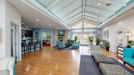 This single-floor community clubhouse is designed to impress from the moment you enter. The centerpiece is a sweeping great room with soaring vaulted ceilings accented by painted teal beams and exposed white rafters. Warm hardwood flooring stretches throughout, while floor-to-ceiling windows and glass doors flood the space with natural light and frame views of an outdoor pool area. A stone-surround fireplace anchors a stylish seating area furnished with teal accent chairs arranged on a circular area rug, creating an inviting focal point. The adjacent entry corridor features bold artwork and additional seating beneath the signature beamed ceiling. The kitchen area is well-appointed with rich dark-wood cabinetry, dark granite countertops, a stainless steel refrigerator, and a breakfast bar with stylish bar seating — ideal for community gatherings. A compact galley-style service kitchen with matching cabinetry provides additional convenience. A cozy lounge off the main hall features plush carpet in deep red, built-in shelving, a tray ceiling, and comfortable seating. The fitness center is equipped with cardio and strength training machines and benefits from natural light through slatted windows. A dedicated business nook with a computer workstation and printer rounds out the amenities of this thoughtfully designed community space.