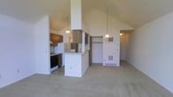This single-floor residence showcases an open-concept layout that seamlessly connects the kitchen and living areas beneath impressively vaulted ceilings that lend a strong sense of height and airiness throughout the space. The kitchen is equipped with dark cabinetry, a gas range, a refrigerator, and a peninsula counter that serves as both a workspace and a casual divider between the cooking and living zones. Pendant lighting hangs from the high ceiling above the peninsula, adding functional illumination and a touch of character to the kitchen area. Adjacent to the kitchen, a built-in shelving nook provides convenient display or storage space. The expansive living area is finished with wall-to-wall carpeting in a neutral tone, creating a warm and comfortable environment. A floor-mounted vent is visible in the living area, indicating a central heating system. The smooth, light-colored walls and vaulted roofline work together to amplify the sense of openness throughout the main living space. A hallway visible toward the rear of the space suggests additional rooms beyond the main open area. The overall ambiance is clean, functional, and welcoming, with the dramatic ceiling height serving as the space's most notable architectural feature.