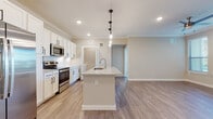 This single-floor unit showcases a thoughtfully designed open-concept layout centered around a well-appointed kitchen. The kitchen is a standout feature, offering crisp white shaker cabinetry with matte black hardware, a tile backsplash, and a full suite of stainless steel appliances including a side-by-side refrigerator with ice and water dispenser, an electric range, and an over-the-range microwave. A generously sized center island with a granite countertop and undermount sink serves as the focal point, illuminated by a trio of pendant lights that hang from the crown-molded ceiling. Recessed lighting and light wood-tone luxury vinyl plank flooring extend throughout the main living areas, lending the space a bright, cohesive feel.

The bedrooms are carpeted for comfort and feature ceiling fans and windows with blinds that invite natural light. Each room offers adequate proportions for comfortable furnishing. The bathroom includes a white vanity with granite countertop, an undermount sink, and a tub-shower combination surrounded by large-format tile. Adjacent to the bathroom, a spacious walk-in closet is equipped with multi-level wire shelving, providing practical and organized storage. Overall, the space presents a clean, neutral palette with modern finishes throughout.