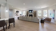 Located at 8134 Colonial Village Dr in Tampa, FL, this single-floor community clubhouse offers a thoughtfully designed open-concept layout that flows seamlessly from one functional zone to the next. The main gathering area is anchored by a sleek fireplace framed by decorative wall art panels, with a wall-mounted flat-screen TV above and crown molding along the ceiling lending a refined architectural touch. Light wood-look flooring runs throughout, complementing the neutral gray walls and recessed lighting that keep the space bright and welcoming. Large windows are positioned throughout the space to invite abundant natural light and provide views of the surrounding greenery. A generous lounge area features plush sectional and accent seating arranged around vibrant area rugs, creating an inviting social atmosphere. Bold, colorful artwork adds energy and personality to the interiors. A dedicated workstation and café-style seating area offer practical spaces for productivity or casual gathering. The layout also includes a kitchen or bar area with pendant lighting and modern appliances, visible through a pass-through opening. Additional seating nooks with varied furniture arrangements provide flexibility for both quiet relaxation and group interaction. The overall ambiance balances contemporary design with a comfortable, community-focused atmosphere ideal for residents.