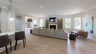 Located at 8134 Colonial Village Dr in Tampa, FL, this single-floor community clubhouse offers a thoughtfully designed open-concept layout that flows seamlessly from one functional zone to the next. The main gathering area is anchored by a sleek fireplace framed by decorative wall art panels, with a wall-mounted flat-screen TV above and crown molding along the ceiling lending a refined architectural touch. Light wood-look flooring runs throughout, complementing the neutral gray walls and recessed lighting that keep the space bright and welcoming. Large windows are positioned throughout the space to invite abundant natural light and provide views of the surrounding greenery. A generous lounge area features plush sectional and accent seating arranged around vibrant area rugs, creating an inviting social atmosphere. Bold, colorful artwork adds energy and personality to the interiors. A dedicated workstation and café-style seating area offer practical spaces for productivity or casual gathering. The layout also includes a kitchen or bar area with pendant lighting and modern appliances, visible through a pass-through opening. Additional seating nooks with varied furniture arrangements provide flexibility for both quiet relaxation and group interaction. The overall ambiance balances contemporary design with a comfortable, community-focused atmosphere ideal for residents.