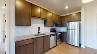 Located at 500 W Broad St in Columbus, Ohio, this single-floor unit showcases a thoughtfully designed contemporary interior with a cohesive mix of warm wood tones, crisp white finishes, and matte black hardware throughout. The open-concept living area features smooth hardwood-style flooring, recessed lighting, and large floor-to-ceiling windows and glass French doors that frame expansive views of the Columbus skyline, filling the space with generous natural light. Pendant lighting accents the transition into the kitchen, which is outfitted with rich dark walnut cabinetry, white subway tile backsplash, light quartz countertops, and a full suite of stainless steel appliances including a refrigerator, range, microwave, and dishwasher. The bedrooms offer plush carpeting and large windows that continue to capture the city views. The bathrooms are finished with floor-to-ceiling white tile, a glass-enclosed walk-in shower, a single undermount sink, quartz countertops, and dark wood cabinetry accented by matte black fixtures. The overall ambiance is clean, modern, and urban, with consistent design details that create a refined and functional living environment in the heart of Columbus.