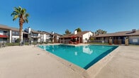 This outdoor amenity space centers around a generously sized rectangular swimming pool with clear, blue-green water, designed to accommodate both leisure swimming and relaxed sunbathing. Depth markers are visible along the pool's edge, indicating a gradually varying depth suitable for a range of swimmers. The pool is enclosed within a wrought iron perimeter fence, providing both security and an open, airy feel.

Surrounding the pool is a broad concrete sun deck outfitted with multiple reclining lounge chairs, offering ample space for sunbathing and outdoor relaxation. A dedicated poolside patio area features a table and chairs shaded by a bold red umbrella, creating a comfortable spot for casual outdoor gatherings. Safety equipment, including a life ring, is mounted near the pool entrance.

The space is framed by mature trees — including a prominent palm tree — that provide natural shade and a lush backdrop against clear blue skies. The surrounding residential buildings feature a traditional architectural style with white exteriors, dark wood trim accents, pitched brown rooflines, and chimney details, contributing to a cohesive and established community aesthetic. Landscaped greenery and potted plants punctuate the pool surround, adding color and texture to the overall environment.