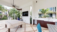 This single-level space centers around a well-appointed covered outdoor living area designed for both relaxation and entertainment. The space features soaring ceilings with clean white stucco finishes, giving it an open and airy feel. A wall-mounted flat-screen TV serves as a focal point, making this area ideal for outdoor viewing and gatherings. A ceiling fan overhead provides comfortable airflow, enhancing the al fresco experience.

The flooring is a standout architectural detail — large-format cream tiles accented with terracotta-toned diamond inlays create a refined, resort-style aesthetic underfoot. Built-in dark mahogany cabinetry with a white countertop offers practical storage and prep space, complementing the overall design with a rich contrast against the white walls.

The space opens to the surrounding environment, with lush tropical foliage — including broad-leafed plants and palms — framing the perimeter and infusing the area with natural, vibrant greenery. Iron railing details provide subtle boundaries while maintaining an open, connected feel to the outdoors. Track and pendant lighting add functional illumination, while the overall layout lends itself to a comfortable, versatile outdoor living experience with a distinctly tropical ambiance.