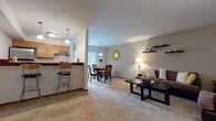 This single-floor residence offers a thoughtfully designed open-concept layout that seamlessly connects the living, dining, and kitchen areas. The living room is carpeted and inviting, featuring vaulted accent walls, floating dark wood shelves, and ample space for relaxed seating. It flows naturally into the dining area, which is anchored by a sliding glass door that fills the space with natural light and provides a view of the greenery outside. The kitchen is a functional galley-style space finished with warm honey-toned cabinetry, dark granite-look countertops, hardwood flooring, and black appliances including a range and refrigerator. A breakfast bar with bar seating connects the kitchen to the main living area, adding convenience and versatility. A secondary room serves well as a den or home office, offering carpet flooring and direct access to the bedroom. The bedroom is a cozy, carpeted retreat with a warm neutral color palette and generous wall space. The bathroom is well-equipped with a single-sink vanity featuring wood cabinetry, hardwood-style flooring, and a full tub-shower combination. Throughout the residence, a consistent warm and neutral color scheme creates a cohesive and comfortable atmosphere.