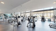 Located at 1010 S Pearl Expy in Dallas, TX, this two-story fitness center offers a comprehensive and thoughtfully designed workout facility. The main floor is anchored by a generously sized open gym area bathed in natural light from floor-to-ceiling windows, which provide views of the surrounding outdoor spaces. This level houses an extensive array of cardio machines—including treadmills, ellipticals, and stair climbers—alongside a wide selection of strength and resistance equipment. Large mirrors line the walls, enhancing the sense of openness throughout the space. A dedicated spin studio on this floor features a full lineup of Spinning bikes, a large wall-mounted display screen, and accessory storage, creating an immersive cycling experience. Adjacent areas provide space for functional and free-weight training, complete with dumbbell racks and benches. A staircase with sleek metal railings connects the main floor to the upper level, which features warm hardwood flooring and a versatile studio space equipped with TRX suspension trainers, resistance tools, stability balls, and mirrored walls—ideal for group classes or individual training sessions. Teal accent walls add a modern, energetic aesthetic throughout, while recessed and pendant lighting complement the abundant daylight. A utility room and closet support day-to-day operations seamlessly.