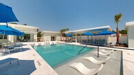 Located at 101 Barnes Ln in Broussard, Louisiana, this single-story outdoor amenity complex delivers a resort-inspired experience designed for relaxation and recreation. At its centerpiece is a generously sized swimming pool with crystal-clear turquoise water, flanked by a broad marble-tiled sundeck. In-pool lounge chairs rest on a submerged shelf, inviting leisurely use directly in the water, while additional chaise lounges and vibrant blue umbrellas are arranged across the deck for comfortable sunbathing.

The surrounding architecture features clean white horizontal siding and a modern aesthetic, with a covered pergola-style structure offering shaded seating along one side of the pool. Black-framed glass doors open seamlessly between indoor and outdoor areas, enhancing the flow of the space. An outdoor television and game area with cornhole boards add a recreational dimension beneath the covered overhang.

Lush tropical landscaping frames the entire complex, with tall palm trees, manicured shrubs, and mulched garden beds providing a vibrant, resort-like atmosphere. A separate patio dining area with a teak table and chairs is nestled among the palms, offering a more intimate outdoor setting. Black iron fencing defines the perimeter, and a turf lawn area provides additional green space. The overall ambiance is bright, open, and polished, evoking a true outdoor retreat.