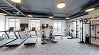 This fitness center offers a comprehensive workout environment designed with both functionality and style in mind. The space features an industrial-modern aesthetic, highlighted by an exposed dark ceiling with visible ductwork and piping, complemented by warm round pendant lighting that creates an inviting atmosphere throughout.

The gym is thoughtfully divided into distinct workout zones. The cardio area lines the windowed wall and includes multiple treadmalls, elliptical trainers, and upright exercise bikes, each equipped with individual display screens for an engaging workout experience. Large windows along multiple walls flood the space with natural light and provide open views of the surrounding exterior.

The strength training section is equally well-appointed, featuring a robust power rack with weight plates, a dual cable functional trainer, a dumbbell rack, and an adjustable weight bench — catering to a wide range of strength and conditioning exercises.

A standout design element is the bold geometric mural on one wall, adding a dynamic visual focal point that elevates the space beyond a typical gym. Soft carpet flooring throughout provides comfort underfoot. A glass-paneled entry door with a dark teal frame adds a refined architectural touch. Wall-mounted TVs are also present, enhancing the overall workout experience in this thoughtfully designed fitness facility.