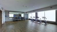 This fitness center offers a well-appointed workout environment designed to support a range of exercise routines. The space features hardwood-style flooring throughout, lending a clean and contemporary feel underfoot. Along one wall, a large floor-to-ceiling mirror visually expands the room and allows users to monitor their form during training sessions. Beneath the mirror, a fully stocked dumbbell rack holds an organized selection of free weights in varying sizes, accompanied by medicine balls and additional accessories. A row of stationary cycling bikes lines the opposite side of the room, positioned directly in front of expansive floor-to-ceiling windows that flood the space with natural light and provide open views to the outside. The bright, airy atmosphere created by the generous window wall enhances the overall workout experience. Recessed lighting in the smooth white ceiling provides additional illumination throughout the space. A centrally located doorway offers access to adjoining areas. The overall design is minimalist and functional, prioritizing open floor space for flexibility, movement, and a variety of training activities in a modern, well-lit setting.