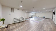 This single-floor yoga and fitness studio offers a generously sized, open-plan space designed to support a variety of wellness activities. The room features durable wood-look flooring throughout, providing a clean, warm aesthetic well-suited to yoga, stretching, and light exercise. Neutral cream-toned walls and recessed lighting create a calm, distraction-free environment conducive to focused practice.

Multiple yoga mats in soft lavender and teal tones are arranged across the floor, accommodating group sessions. Fitness accessories, including stability balls and yoga blocks, are available within the space. A wall-mounted flat-screen television is positioned on one wall, enabling guided workout sessions or instructional content.

Natural light enters through several windows fitted with horizontal blinds, offering views of the surrounding exterior while allowing light control. A wall-mounted PTAC unit provides independent climate control, ensuring comfort year-round. A multi-tier metal shelving rack near the entrance provides convenient storage for personal belongings or equipment.

The studio also includes a dedicated smaller alcove area, suitable for individual practice or one-on-one instruction. Overall, the space presents a clean, functional, and welcoming environment tailored to yoga and general fitness use.