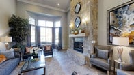 This single-story clubhouse offers a sophisticated and welcoming gathering space designed with upscale finishes and thoughtful details throughout. The centerpiece of the main lounge is a striking floor-to-ceiling stone fireplace, clad in natural stacked stone and adorned with decorative mirrors and wall sconces that add warmth and visual interest. The soaring ceiling height amplifies the grandeur of the room, creating an open and airy atmosphere. Large bay windows along one wall allow abundant natural light to pour in, offering views of the surrounding outdoor greenery and making the space feel connected to the exterior landscape. The flooring features large-format tile in neutral tones, complementing the room's refined color palette of warm grays and taupe. Seating areas are furnished with stylish leather chairs and sofas accented with decorative pillows, providing comfortable spaces for relaxation or socializing. Curtain-draped windows and carefully placed floral arrangements add elegant finishing touches. Overall, the clubhouse strikes a balance between sophisticated design and inviting comfort, making it an ideal communal retreat.