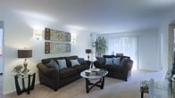This single-floor space offers a thoughtfully designed living area that combines comfort and contemporary style. The living room serves as the centerpiece, featuring plush dark chocolate-toned sofas accented with teal and patterned throw pillows, creating a rich contrast against the light neutral carpeting and crisp white walls. A round glass-top coffee table anchors the seating arrangement, adding a touch of elegance to the space.

Natural light pours in through large sliding glass doors dressed with sheer white vertical blinds, brightening the entire room and creating an airy, open ambiance. Wall-mounted sconce lighting adds a warm, decorative glow alongside a classic table lamp, ensuring the space is well-lit at any time of day.

Decorative elements are carefully curated throughout — a set of framed botanical wall art brings visual interest to one wall, while a sculptural bamboo accent and decorative branches add organic texture and depth to the room. The smooth, light-colored ceiling enhances the sense of openness overhead.

Overall, this space presents a clean, inviting atmosphere with a cohesive design palette that balances modern furnishings with natural accents, making it a comfortable and visually appealing environment for everyday living.