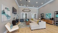 Located at 2889 Sollie Rd in Mobile, Alabama, this thoughtfully designed single-floor space combines sophisticated style with an open, airy layout. The expansive living area immediately draws attention with its soaring tray ceilings, crown molding, and a striking circular chandelier that anchors the room. Light hardwood flooring flows throughout, complementing the soft gray walls and white trim for a clean, contemporary aesthetic. Large French doors flood the space with natural light and provide seamless access to the outdoors. A generously sized sectional sofa, accent chairs, and curated artwork create a warm, inviting atmosphere. Adjacent to the living area, a refined lounge or sitting nook features textured accent wallpaper, open metal shelving with decorative objects, and an intimate bistro-style seating arrangement. The kitchen area is a functional centerpiece, boasting a large quartz-topped island with bar seating, rich dark wood cabinetry, a double sink, and a row of amber glass pendant lights that add a warm, elegant glow. Stainless steel appliances and abundant natural light from multiple windows complete the kitchen's upscale appeal. A billiard table visible in the open floor plan adds a recreational dimension to this versatile space. Every detail reflects a cohesive, upscale design sensibility throughout.