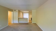 This single-floor residence offers a practical and functional open-concept layout that seamlessly connects the living area to the kitchen. The living room is finished with wall-to-wall carpeting in a neutral tone, creating a warm and comfortable atmosphere throughout the space. Smooth, light-colored walls complement the overall bright and airy feel of the interior.

The kitchen is defined by a peninsula counter that serves as a natural divider between the living and cooking areas, offering both workspace and a casual interaction point. The kitchen is equipped with standard appliances including a range and refrigerator, and features tile or hard flooring that contrasts with the carpeted living space. A window dressed with vertical blinds allows natural light to filter into the kitchen and adjacent areas, enhancing the openness of the layout.

The textured ceiling adds a subtle visual detail throughout the unit. A sliding closet with bifold doors is visible near the entryway, providing convenient storage. The neutral color palette of warm beige and off-white tones used throughout the walls and finishes gives the space a clean, cohesive appearance. Overall, the layout is efficient and well-suited for everyday living.