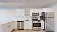 This thoughtfully designed single-floor unit offers a bright, modern living experience with a cohesive aesthetic carried throughout every room. Light-toned luxury vinyl plank flooring runs continuously across the space, complementing the crisp white walls and recessed lighting that keep the interior feeling open and airy.

The kitchen is a standout feature, outfitted with white shaker-style cabinetry, open shelving, and light quartz countertops. A full suite of stainless steel appliances includes a gas range, over-the-range microwave, dishwasher, and refrigerator. A single-basin undermount sink with a modern arc faucet completes the functional workspace. The kitchen flows naturally into the living area, which benefits from a large window fitted with vertical blinds, allowing for ample natural light and a connection to the outside.

The bathroom maintains the same clean, white palette with a vanity featuring shaker-style cabinetry, a white countertop, an undermount sink, and a lighted mirror above. Light wood-tone flooring continues here, adding warmth to the space.

A dedicated closet with a sliding barn-style door provides practical storage. Exposed beam detailing on the ceiling adds a subtle architectural touch, while recessed lighting throughout ensures the entire unit is well-illuminated. The overall ambiance is modern, clean, and move-in ready.