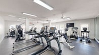 Located at 5489 Crescent Ridge Dr in Dublin, Ohio, this well-appointed fitness center spans a generous single floor divided into four distinct workout areas, offering a comprehensive environment for exercise and wellness. The main cardio zone is bright and open, lined with a row of True treadmills and elliptical machines, all positioned beneath flush skylights and ceiling fans that ensure consistent airflow and visibility. Full-length mirrored walls expand the sense of space and allow users to monitor their form throughout their workout. A dedicated strength training room features green-tiled walls, large windows overlooking lush outdoor greenery, a power rack with barbells, a dumbbell rack, and a wall-mounted TV — creating a focused environment for resistance training. Additional areas include a multi-use exercise space equipped with a cable machine and functional training equipment, as well as a transitional lounge or utility room that provides a comfortable break area. Throughout the facility, white-painted walls and columns, durable dark rubber flooring, and multiple ceiling fans contribute to a clean, functional aesthetic. Natural light flows in from multiple windows and glass-paneled doors, keeping the atmosphere bright and energizing. The layout is thoughtfully organized to support a full range of fitness activities within a single, cohesive facility.
