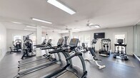 Located at 5489 Crescent Ridge Dr in Dublin, Ohio, this well-appointed fitness center spans a generous single floor divided into four distinct workout areas, offering a comprehensive environment for exercise and wellness. The main cardio zone is bright and open, lined with a row of True treadmills and elliptical machines, all positioned beneath flush skylights and ceiling fans that ensure consistent airflow and visibility. Full-length mirrored walls expand the sense of space and allow users to monitor their form throughout their workout. A dedicated strength training room features green-tiled walls, large windows overlooking lush outdoor greenery, a power rack with barbells, a dumbbell rack, and a wall-mounted TV — creating a focused environment for resistance training. Additional areas include a multi-use exercise space equipped with a cable machine and functional training equipment, as well as a transitional lounge or utility room that provides a comfortable break area. Throughout the facility, white-painted walls and columns, durable dark rubber flooring, and multiple ceiling fans contribute to a clean, functional aesthetic. Natural light flows in from multiple windows and glass-paneled doors, keeping the atmosphere bright and energizing. The layout is thoughtfully organized to support a full range of fitness activities within a single, cohesive facility.