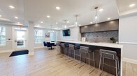 Located at 1 E Pearl St in Burlington, NJ, this single-floor community amenity space is designed to offer residents a sophisticated and versatile gathering environment. The open-concept layout is anchored by a spacious central lounge featuring tray ceilings with recessed lighting, a wall-mounted fireplace beneath a flat-screen TV, and a coordinated seating arrangement of sleek black leather sofas and armchairs set atop patterned area rugs over light wood-look tile flooring. Large windows allow natural light to flow throughout, enhancing the bright and airy atmosphere.

A prominent wet bar runs along one side of the space, finished with dark wood cabinetry, a textured stone backsplash, white quartz countertops, and pendant glass lighting above a full row of modern barstools — ideal for social gatherings. Adjacent to the bar, a dedicated dining area features a round table with navy blue chairs beneath a classic chandelier.

The game room, finished in soft blue-gray tones with wainscoting and patterned carpet, accommodates a billiards table and a foosball table alongside high-top tables and seating. Together, these areas create a well-appointed, multi-functional community space suited for both relaxation and entertainment.