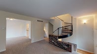 This three-floor residence is defined by its striking architectural centerpiece: a curved spiral staircase with black metal railings, anchored by a decorative brick base that adds a classic, structural character to the main living area. The living space is generously proportioned, finished with neutral-toned wall-to-wall carpeting and painted in warm, light neutral tones throughout, creating a cohesive and inviting atmosphere. Natural light filters in through multiple windows fitted with horizontal blinds, brightening each room.

The galley-style kitchen is a functional and well-organized space, featuring white cabinetry, dark countertops, a tile backsplash, and hardwood flooring. It is equipped with a full suite of white appliances, including a range and refrigerator, with ample upper and lower cabinet storage lining both sides of the corridor-style layout.

The bathroom includes a tub-shower combination with white tile surround, a large vanity mirror with overhead lighting, a single-basin sink set into a dark granite-style countertop, and wood-tone flooring.

The multiple bedrooms across the floors are carpeted and neutrally finished, each offering closet access and natural light from windows. Wall sconces in select rooms provide warm accent lighting, contributing to the overall comfortable and functional character of this well-structured multi-level home.