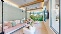 Located at 1464 Laurel Sound Lane in Sanford, Florida, this thoughtfully designed single-floor community space offers a welcoming and resort-inspired atmosphere. The open-plan layout is anchored by wide-plank light hardwood flooring that flows throughout the main gathering area, complemented by recessed lighting and clean white walls that create a bright, airy ambiance.

A standout architectural feature is the dedicated lounge alcove, where rope-suspended swing beds hang from exposed wood-beam ceilings with warm accent lighting. Shiplap walls in a soft sage green lend a coastal character to this zone, while light brick-style tile flooring adds textural contrast. A vivid hand-painted tropical mural depicting lush palm trees and a serene waterscape serves as a dramatic focal point, reinforcing the Florida-inspired design narrative.

The main social area accommodates multiple seating arrangements, including woven rattan chairs and neutral upholstered sofas, making it ideal for both casual gatherings and relaxed lounging. Large floor-to-ceiling glass panels and sliding doors flood the space with natural light and offer direct views of an outdoor pool area, seamlessly blending the interior with the surrounding environment.

Overall, the space presents a cohesive tropical-coastal aesthetic, balancing playful design elements with comfortable, functional communal living.