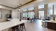 Located at 1800 St. Joseph Pkwy in Houston, TX, this refined two-floor amenity space offers a generous and thoughtfully designed layout spread evenly across both levels. The ground floor makes an immediate impression with soaring double-height ceilings, large-format tile flooring in a herringbone pattern, and dramatic floor-to-ceiling windows that flood the space with natural light and provide views of an outdoor pool and landscaped surroundings. A sleek wet bar with pendant lighting and bar seating anchors the open-plan living and dining area, which accommodates both communal dining and lounge seating. Rich wood paneling with a wall-mounted display screen adds warmth and a contemporary focal point to the main living room. An elegant staircase with ornate iron balusters and a warm wood handrail connects the two floors, framed by a striking textured mosaic accent wall. The upper level continues the refined aesthetic with a quieter lounge area featuring built-in bookshelves, a mirrored vanity console framed in metallic mosaic tile, and plush seating. Soft carpet, curated artwork, and carefully selected furnishings throughout create a warm, upscale ambiance suited for both relaxation and social gathering.