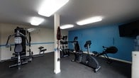 This fitness space is designed to support a well-rounded workout routine within a compact, efficiently arranged single-floor layout. The room is defined by crisp white walls accented by a bold blue feature wall, creating an energetic and motivating atmosphere. Bright flush-mounted ceiling panels provide consistent, even illumination throughout the space, ensuring excellent visibility across all workout areas.

The flooring consists of durable, dark-toned material well-suited for high-traffic exercise use. A central structural column divides the space while resistance bands and accessories are neatly mounted to it, making practical use of the vertical surface.

The equipment selection covers a range of fitness needs. Cardio options include a recumbent stationary bike and a rowing machine, both positioned toward the center and side of the room for easy access. A multi-function cable and pulley strength training machine occupies a dedicated area, while an additional piece of strength equipment is visible near the far wall. The layout allows users to move between stations with reasonable ease.

Overall, the space offers a functional and organized fitness environment with a clean, purposeful aesthetic suitable for everyday training.