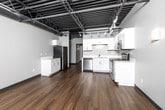 Located at 660 E Fifth St in Des Moines, Iowa, this one-bedroom, one-bathroom unit delivers a striking industrial-modern aesthetic throughout. The open-concept kitchen and living area serves as the heart of the space, showcasing soaring ceilings with exposed steel trusses, ductwork, and dark structural beams that lend a distinctive loft-style character. Track lighting and ceiling fans complement the airy volume overhead, while warm hardwood flooring runs continuously throughout, grounding the industrial design with natural warmth.

The kitchen is outfitted with white cabinetry, light countertops, and a full suite of stainless steel appliances including a refrigerator, range, built-in microwave, and dishwasher. The living area benefits from direct outdoor access through a glass-paneled door, flanked by clerestory windows that draw in generous natural light and views of the surrounding greenery.

The spacious bedroom continues the exposed ceiling design and features its own door to the outdoors, an additional window for cross-ventilation, and direct access to a dedicated walk-in closet equipped with wire shelving systems. The full bathroom offers a tub-shower combination, toilet, and vanity with hardwood-style flooring tying seamlessly into the rest of the unit.