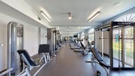 Located at 497 Angliana Ave in Lexington, Kentucky, this fitness center offers a comprehensive, single-floor exercise facility designed to accommodate a wide range of workout needs. The gym is generously proportioned, with a long, open layout that provides ample space between equipment stations for comfortable use.

The cardio section is prominently featured along one wall, lined with multiple treadmills, elliptical machines, and stationary bikes. Large arched windows and full-height glass doors along this side of the room flood the space with natural light and offer views of the outdoor greenery beyond, creating an energizing workout environment. Ceiling fans and recessed linear lighting complement the bright, airy atmosphere throughout.

The strength training area is equally well-appointed, featuring a multi-functional Jones machine power rack, adjustable benches, a full dumbbell rack with a wide range of weights, and various selectorized cable and resistance machines. Wall-mounted mirrors extend across key sections of the gym, enhancing the sense of space and allowing for form monitoring during exercise. A mounted flat-screen television adds to the amenity.

Adjacent to the main fitness floor, a dedicated package locker room provides secure parcel storage, accessed through a clean entryway with speckled flooring, warm wood-trimmed doors, and a welcoming bench — adding a practical and polished touch to the overall facility.
