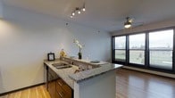 This single-floor space showcases a thoughtfully designed open-concept layout that seamlessly connects the kitchen and living areas. The kitchen is a standout feature, highlighted by a generous granite-topped peninsula that doubles as a breakfast bar, complete with a stainless steel sink and dark cabinetry offering ample storage. Track lighting overhead provides focused illumination throughout the kitchen area, while a ceiling fan in the adjacent living space promotes comfortable air circulation year-round. The warm hardwood flooring runs throughout, lending a cohesive and inviting feel to the entire space. A large window with horizontal blinds floods the living area with natural light, offering views of the surrounding greenery outside. The clean, neutral wall tones complement the granite surfaces and dark wood cabinetry, creating a balanced and contemporary aesthetic. The open layout encourages a natural flow between cooking, dining, and living areas, making the space both functional and welcoming. Overall, the unit combines modern finishes with practical design, resulting in a bright and comfortable living environment.