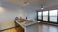 This single-floor space showcases a thoughtfully designed open-concept layout that seamlessly connects the kitchen and living areas. The kitchen is a standout feature, highlighted by a generous granite-topped peninsula that doubles as a breakfast bar, complete with a stainless steel sink and dark cabinetry offering ample storage. Track lighting overhead provides focused illumination throughout the kitchen area, while a ceiling fan in the adjacent living space promotes comfortable air circulation year-round. The warm hardwood flooring runs throughout, lending a cohesive and inviting feel to the entire space. A large window with horizontal blinds floods the living area with natural light, offering views of the surrounding greenery outside. The clean, neutral wall tones complement the granite surfaces and dark wood cabinetry, creating a balanced and contemporary aesthetic. The open layout encourages a natural flow between cooking, dining, and living areas, making the space both functional and welcoming. Overall, the unit combines modern finishes with practical design, resulting in a bright and comfortable living environment.