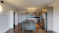 Located at 951 Government St in Mobile, Alabama, this single-floor unit offers a sleek, modern aesthetic defined by polished concrete floors that flow seamlessly throughout the entire space. The open-concept kitchen serves as a focal point, outfitted with wood-toned cabinetry extending to the ceiling for ample storage, white quartz countertops, a dark tile backsplash, and a full suite of stainless steel appliances including a refrigerator, range, microwave, and dishwasher. A mobile kitchen island with an industrial metal finish adds flexible prep and serving space. The living area is bright and airy, bathed in natural light from large windows that frame views of the surrounding Mobile cityscape. The bedroom is a calm, neutral retreat featuring wall-mounted sconce lighting and direct access to a well-appointed bathroom. The bathroom includes a vessel sink set atop a wood-finish vanity with ample counter space, a large mirror, and a glass-enclosed tile shower. Throughout the space, a consistent modern design language is maintained through clean lines, neutral tones, and thoughtful material choices, creating a cohesive and contemporary urban living environment.