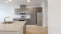 Located at The Carlyle at Chatham Hills in Westfield, Indiana, this two-bedroom, two-bathroom residence offers a thoughtfully designed single-level layout with contemporary finishes throughout. The open-concept kitchen and living area serves as the heart of the home, showcasing grey shaker-style cabinetry, granite countertops, a subway tile backsplash, stainless steel appliances, and a central island with an undermount sink — all set atop light-toned hardwood-style flooring. Pendant lighting adds a refined touch above the island. The living area flows naturally from the kitchen, benefiting from large windows that invite abundant natural light and provide views of the surrounding greenery. Both bedrooms feature plush carpeting and windows overlooking outdoor scenery, creating comfortable and well-lit retreats. The two bathrooms are finished with granite-topped vanities and dark cabinetry. The full bathroom includes a tiled tub-shower combination, while the second bathroom offers a clean, functional layout. A dedicated closet provides additional storage. Neutral grey tones and clean white trim throughout the residence create a cohesive, modern aesthetic that is both inviting and versatile.