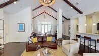 This single-story clubhouse is defined by its dramatic vaulted ceilings adorned with dark exposed wood beams, creating an impressive architectural focal point throughout the space. The open-concept layout flows seamlessly from one area to the next, all unified by rich, dark hardwood flooring and crisp white walls that lend a clean, welcoming atmosphere.

The main lounge area serves as the heart of the clubhouse, furnished with a substantial leather sofa accented by colorful throw pillows and complemented by neutral seating. Statement chandeliers—including a geometric orb fixture and a starburst-style pendant—add character and warmth. A dedicated media area features a wall-mounted flat-screen television flanked by open shelving units, ideal for relaxed gatherings.

A kitchen bar with dark cabinetry, pendant lighting, and counter-height seating provides a functional space for socializing, while a casual dining area with a round pedestal table and workstation nook adds versatility. Floor-to-ceiling windows and glass double doors along one wall flood the interior with natural light and offer direct views and access to an outdoor pool area, making the transition between indoor and outdoor living effortless.