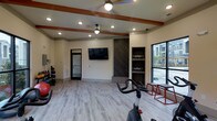 This well-appointed fitness center offers a clean, modern environment designed to support a variety of workout routines. The space features light wood-style flooring throughout, creating a bright and welcoming atmosphere that complements the neutral wall tones. Decorative wood ceiling beams add a warm architectural touch, while recessed lighting and dual ceiling fans ensure the space remains comfortable and well-lit during any workout session.

The main workout area is open and spacious, accommodating cardio equipment including a stationary cycle, as well as strength training tools such as free weights and weighted platforms. Built-in shelving with a dark wood accent surround provides organized storage for fitness accessories, flanked by a wall-mounted flat-screen television for entertainment during workouts.

Large windows along one wall allow natural light to pour in while offering views of the surrounding outdoor area, enhancing the overall sense of openness. A glass-paned interior door connects to an adjacent room, extending the functional layout of the facility. The overall design balances practicality with aesthetic appeal, making this fitness center a polished and fully equipped space for everyday exercise needs.