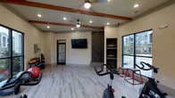 This well-appointed fitness center offers a clean, modern environment designed to support a variety of workout routines. The space features light wood-style flooring throughout, creating a bright and welcoming atmosphere that complements the neutral wall tones. Decorative wood ceiling beams add a warm architectural touch, while recessed lighting and dual ceiling fans ensure the space remains comfortable and well-lit during any workout session.

The main workout area is open and spacious, accommodating cardio equipment including a stationary cycle, as well as strength training tools such as free weights and weighted platforms. Built-in shelving with a dark wood accent surround provides organized storage for fitness accessories, flanked by a wall-mounted flat-screen television for entertainment during workouts.

Large windows along one wall allow natural light to pour in while offering views of the surrounding outdoor area, enhancing the overall sense of openness. A glass-paned interior door connects to an adjacent room, extending the functional layout of the facility. The overall design balances practicality with aesthetic appeal, making this fitness center a polished and fully equipped space for everyday exercise needs.