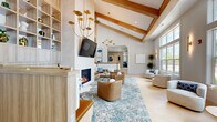 Located at 1153 Bees Ferry Rd on Johns Island, SC, this single-floor community clubhouse offers a thoughtfully designed, open-concept layout that blends coastal sophistication with warm, modern interiors. The space is immediately distinguished by its dramatic vaulted ceilings accented with exposed natural wood beams, which add architectural character and a sense of grandeur throughout.

The living area serves as the heart of the space, centered around a sleek floor-to-ceiling fireplace flanked by built-in light wood shelving with integrated bench seating. A wall-mounted display and a statement gold chandelier complete the focal point. Comfortable seating arrangements, including leather swivel chairs and neutral-toned sofas atop a teal patterned area rug, create an inviting communal atmosphere.

Adjacent to the lounge, a wall of oversized windows floods the space with abundant natural light while offering lush views of the surrounding palm-dotted landscape. A secondary seating area with rounded upholstered chairs takes full advantage of this sunlit corner.

The kitchen and bar area features an extended marble-topped island with hex-tile detailing, light wood cabinetry, white quartz countertops, gold fixtures, and open shelving that extends to the ceiling. A coffee station, beverage refrigerator, and white tile backsplash round out this well-appointed amenity space. Light hexagonal tile flooring flows seamlessly throughout, tying every zone together cohesively.