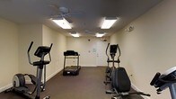 This fitness room is a dedicated exercise space designed to support cardio-focused workouts. The room features dark carpeted flooring and light-colored walls, creating a clean, functional environment. Overhead, two ceiling fans with integrated light fixtures work alongside flush-mounted fluorescent panel lights to ensure the space remains well-lit and properly ventilated during workouts. The equipment selection includes multiple treadmills, an elliptical trainer, and a stationary bike, all positioned to allow comfortable use without overcrowding. A standard door with an illuminated exit sign provides clear egress, and a smoke detector is visible on the ceiling, highlighting attention to safety. The layout keeps the space straightforward and easy to navigate, with outlets along the walls offering convenient access for personal devices. While the room is compact, the arrangement of equipment is practical and efficient, making it a suitable amenity for regular cardiovascular exercise.