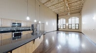Located in downtown Flint at 540 S Saginaw Street, this two-bedroom, two-bathroom two-story unit showcases an impressive industrial-loft aesthetic rooted in thoughtful architectural design. The main level is anchored by a generous living area with rich, dark hardwood floors that gleam under natural light pouring through striking arched windows. These oversized windows frame panoramic views of the surrounding downtown cityscape, lending the space a distinctly urban character. The soaring exposed ceiling with visible structural trusses and ductwork reinforces the loft-style industrial charm throughout the main floor. A staircase with a modern cable railing and wood handrail connects the two levels seamlessly. The kitchen is situated centrally on the main floor, complementing the open-concept flow. Both bathrooms are finished with tile flooring; one features a dark-stained vanity with a white countertop, while the other includes a light wood vanity with dark granite countertop and a walk-in shower. The upper level, accessed via the staircase, houses the expansive primary bedroom and a second carpeted bedroom, both beneath the exposed truss ceiling system. Together, the two floors offer a distinctive, loft-inspired living experience in the heart of downtown Flint.