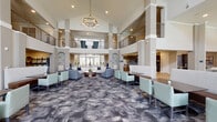 Located at 25500 Westheimer Pkwy in Katy, Texas, this elegant community clubhouse is designed to impress from the moment you enter. The grand lobby commands attention with its soaring double-height ceilings, an upper mezzanine level framed by sleek railings, and decorative columns that lend a sense of classical sophistication. Multiple seating arrangements, featuring curved cream sofas, navy accent chairs, and patterned area rugs, create inviting social spaces throughout the open floor plan. Floor-to-ceiling windows flood the interior with natural light and offer direct views of the resort-style pool area beyond. A pair of plush teal wingback chairs positioned before the expansive windows makes for a particularly striking vantage point. The dining area is anchored by a striking circular chandelier and features geometric parquet-style flooring, dark wood furnishings, and a built-in service counter with a tile backsplash. A fully appointed communal kitchen showcases dark cabinetry, a large center island with light stone countertops, stainless steel appliances, and bold navy pendant lighting. Connecting hallways with tasteful artwork and herringbone-patterned flooring tie the spaces together seamlessly, creating a cohesive, upscale environment ideal for community gatherings and everyday relaxation.