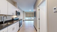 Located at 225 Innovative Way in Scarborough, Maine, this efficiently designed single-floor unit offers a clean, contemporary aesthetic throughout its compact footprint. Light hardwood flooring runs continuously across all areas, creating a cohesive and airy feel. The galley-style kitchen is outfitted with white shaker cabinetry featuring brushed nickel hardware, dark granite countertops, a stainless steel sink, and a full suite of stainless steel appliances including a refrigerator, electric range, and over-the-range microwave. An entry corridor provides a welcoming transition into the unit, with a keypad-secured front door and a wall-mounted thermostat control panel. The living area sits at the far end of the unit, bathed in natural light from a wide, multi-panel window dressed with neutral roller shades that offer views of the surrounding streetscape. Neutral gray and warm beige wall tones complement the light flooring throughout. The bathroom features a white vanity with dark granite countertop, an undermount sink with brushed nickel fixtures, a large mirror, and coordinating hardware accents. The unit's modern, minimalist design and functional layout make it a practical and well-appointed space in Scarborough.