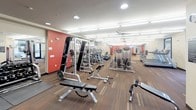 This fitness center is a thoughtfully designed, single-floor facility that offers a comprehensive workout environment for users of all fitness levels. The space is organized into distinct functional zones, each tailored to a specific type of training. The main floor features patterned carpet flooring throughout, recessed and track lighting, and ceiling fans that contribute to a comfortable exercise environment.

The strength training area is well-appointed with free weights, a full dumbbell rack, multiple weight benches, and cable machines — including a prominent Precor-brand unit. A bold red accent wall adds energy to this zone, which also benefits from mirrored walls that enhance the sense of space. Nearby, a dedicated cardio section houses a row of treadmills, elliptical trainers, and stair climbers, with large windows that bring in natural light and views of the surrounding exterior.

A glass-walled studio provides a semi-private space for cycling workouts, complete with stationary bikes and a wall-mounted display screen. Adjacent to the main workout floor, a welcoming entry area includes a beverage cooler, cubbies for personal storage, a small-screen thermostat panel, wall art, and a waste receptacle — offering convenient amenities before and after workouts. Multiple mounted televisions throughout the facility ensure entertainment is always within view during training sessions.