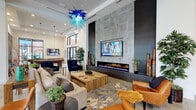 Located at 2629 Wagon Wheel Rd in Oxnard, California, this single-story community clubhouse showcases contemporary design with an upscale, resort-inspired ambiance. The main lounge area is anchored by a dramatic floor-to-ceiling stone-clad fireplace wall with an integrated flat-screen TV and a sleek linear electric fireplace, all set beneath soaring ceilings adorned with a striking blue blown-glass chandelier. Plush seating arrangements, including a sectional sofa, navy velvet accent chairs, and cognac leather chairs, create an inviting and layered social atmosphere. Large-format tile flooring flows throughout the space, lending a cohesive, polished look. A dedicated game area features a billiard table and a built-in dark cabinetry wall with a tufted bench and decorative metalwork. The dining area offers multiple seating configurations alongside floor-to-ceiling steel-framed windows that invite natural light and outdoor views. A vivid marina mural serves as a focal point in the communal dining and kitchen space, which includes a long communal high-top table, a stainless steel refrigerator, and a well-appointed wet bar with dark cabinetry, quartz countertops, and a textured wave-tile backsplash. Curated artwork and lush indoor plants complete the sophisticated, welcoming environment.
