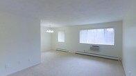 This single-floor space presents a clean, functional layout designed for comfortable everyday living. The main living area is generously proportioned, featuring plush wall-to-wall carpeting that adds warmth underfoot and contributes to the room's quiet, comfortable atmosphere. Crisp white walls and a bright, neutral color palette throughout create an open and airy feel, maximizing the sense of space.

Natural light flows in through multiple windows dressed with vertical blinds, including a large picture window that serves as a focal point of the main room. A window-mounted air conditioning unit provides convenient climate control during warmer months, while baseboard heaters along the walls ensure efficient warmth in cooler seasons.

A classic chandelier with a multi-arm design adds a touch of elegance to the dining or living area, serving as a subtle decorative accent against the otherwise understated interior. The open-plan configuration between the living and dining spaces allows for flexible furniture arrangements and easy movement throughout.

Overall, this space offers a bright, well-lit environment with practical heating and cooling solutions, neutral finishes, and an inviting layout suited to a variety of lifestyles.