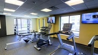 This fitness center is designed to support a well-rounded workout routine within a compact, efficiently organized space. The room features a drop ceiling with recessed panel lighting that provides consistent illumination throughout. Large windows along two walls allow ample natural light to filter in, offering views of the surrounding urban environment and creating an open, airy atmosphere despite the room's focused footprint.

The flooring consists of durable rubber matting, a practical choice that provides cushioning and stability for various types of exercise. The walls are painted in a bold yellow accent, lending the space an energetic and motivating ambiance.

Equipment includes Matrix-branded cardio machines—a treadmill, stationary bike, and rowing machine—positioned to take advantage of the natural light near the windows. A multi-tiered dumbbell rack holds a full range of free weights, accompanied by an additional vertical weight storage tower, providing options for strength training. A wall-mounted flat-screen television is centrally positioned, allowing users on any machine to view it comfortably. The overall layout is thoughtfully arranged to maximize usability while maintaining clear pathways between equipment.