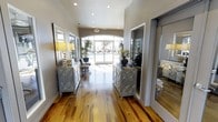 This single-story space makes a strong first impression through its well-appointed entryway corridor. Rich, variegated hardwood flooring in warm honey and caramel tones runs the length of the hallway, adding natural warmth to the interior. The walls are finished in a contemporary warm gray, providing a neutral backdrop that complements the refined décor.

Flanking the corridor are two matching mirrored and metallic console tables, each styled with decorative lamps featuring yellow shades and lush tropical potted plants, lending an upscale, curated feel to the space. Large floor-length mirrors are positioned along the walls, amplifying the sense of depth and light throughout.

Recessed lighting in the ceiling provides even, ambient illumination, while a generous arched window at the far end draws in abundant natural light and frames a view of an outdoor pool area, creating a bright and inviting focal point. The overall ambiance is polished and welcoming, blending contemporary design elements with warm natural materials to create a space that feels both elegant and functional.