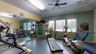 This fitness center offers a comprehensive workout environment within a compact, efficiently organized single-room layout. The space features durable rubber flooring throughout, providing a safe and practical surface for exercise. Two ceiling fans ensure steady air circulation, complemented by overhead fluorescent lighting that keeps the room consistently well-lit.

The cardio zone, situated along the windowed wall, includes treadmills, an elliptical trainer, and upright and recumbent stationary bikes. A row of windows along one wall allows natural light to filter in and offers views of an outdoor pool area, creating an inviting atmosphere during workouts. A wall-mounted television is positioned for easy viewing from the cardio equipment.

A full-wall mirror on one side of the room visually expands the space and is flanked by resistance bands and stability balls, forming a dedicated functional training area. A multi-station cable and weight machine anchors the strength training section, offering a variety of resistance exercises. Additional equipment, including a leg press and bench, rounds out the strength offerings.

Informational boards, a bulletin board, and posted fitness center rules contribute to the organized, community-oriented character of the space. The two-toned walls — warm beige on the upper half and muted teal-green on the lower — give the room a clean, purposeful aesthetic suited to an active fitness environment.