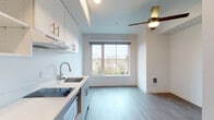 Located at 105 N Killingsworth St in Portland, Oregon, this single-floor unit offers a clean, modern design with an efficient and well-appointed layout. The open living area is finished with light wood-tone luxury vinyl plank flooring throughout, creating a cohesive and contemporary feel. A ceiling fan provides comfort and adds a functional accent to the main living space.

The kitchen is thoughtfully designed with two-tone cabinetry — crisp white upper cabinets paired with soft grey lower cabinets — complemented by light countertops, a stainless steel sink with a pull-down faucet, and built-in appliances including a cooktop, microwave, and refrigerator. Ample upper cabinet storage and a range hood complete this practical cooking space.

Large windows in both the kitchen and main living area allow generous natural light to fill the unit while offering views of the surrounding Portland neighborhood. The bathroom features a clean, minimalist design with accessibility-friendly fixtures, including a grab bar for added safety and convenience.

The unit is accessed through a solid-core entry door, and the layout, as visible in the dollhouse view, provides a well-organized flow between the kitchen, living area, bathroom, and entry corridor. Overall, the space presents a bright, functional, and move-in-ready environment in North Portland.