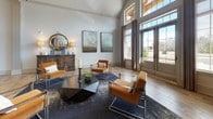 Located at 100 Whitworth Way in Williamsburg, Virginia, this single-story community amenity center offers a well-rounded collection of spaces designed for both relaxation and active living. The welcoming lounge area immediately sets the tone with soaring vaulted ceilings, warm hardwood-style flooring, and an abundance of natural light streaming through tall, floor-to-ceiling windows and double French doors. Contemporary amber leather accent chairs, a geometric coffee table, and curated landscape artwork complete the sophisticated yet inviting atmosphere. Adjacent to the lounge, a fully appointed kitchen and social area features sleek grey cabinetry, a large marble-topped island with a built-in sink, wood-look tile flooring, and a large wall-mounted television — ideal for community gatherings and events. The fitness facilities are extensive and thoughtfully divided. A dedicated cardio and strength training room is equipped with treadmills, cycling bikes, cable machines, and rubber flooring, with natural light provided by multiple windows. A separate yoga and group exercise studio offers light hardwood floors, floor-to-ceiling mirrors, a large display screen, exercise mats, kettlebells, and stability balls, creating an ideal environment for group classes or individual practice. Convenient amenities such as dual water fountains with a bottle-filling station are also present throughout the facility.