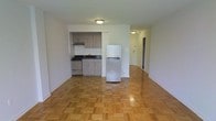 This single-floor unit presents an open-concept layout that combines the living area and kitchen into one cohesive space. The kitchen is neatly tucked into a recessed alcove, fitted with light wood cabinetry, a gas range with a stainless steel finish, a double-basin sink with a dark countertop, and a full-size refrigerator that stands at the boundary between the kitchen and the living area. Under-cabinet space and upper cabinetry provide practical storage. The living area is spacious and bright, featuring classic parquet hardwood flooring laid in a basket-weave pattern that adds warmth and texture underfoot. Neutral white walls keep the interior feeling clean and open. Flush-mount ceiling lighting illuminates both the main living space and a hallway visible in the background, which leads to additional rooms. The hallway suggests further private spaces beyond the main area. Overall, the unit offers a straightforward, functional design with timeless flooring details and a practical kitchen setup suited for everyday living.